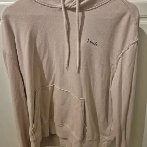 Beige hoodie från H&M Divided - Säljer en beige hoodie från H&M Divided med broderad text 'Smile' på bröstet. Tröjan har huva med snörning, magficka och långa ärmar. Perfekt för en chill och avslappnad stil. Mjuk insida i bomullsblandning.