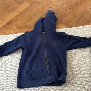 Marinblå hoodie från Polo Ralph Lauren - Säljer en marinblå hoodie från Polo Ralph Lauren med dragkedja och huva. Tröjan har en klassisk röd logga broderad på bröstet, känguruficka framtill och ribbade muddar. Perfekt för chill dagar eller när du vill ha en clean och stilren look.