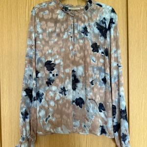 Blus från InWear - Tunn beige blå polyesterblus från InWear. Volangdetaljer i krage och ärmslut. En knapp i halskragen men är lika snygg oknäppt. Endast använd en gång.