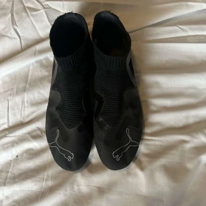 Svarta fotbollsskor från Puma - Snygga svarta fotbollsskor från Puma med vit logga och detaljer. Skorna har strumpliknande skaft i stickad syntet och plösfri design för tight passform. Yttersulan har fasta dobbar för bra grepp på planen. Perfekta för dig som vill sticka ut på matchen.