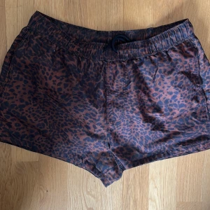 Leopardmönstrade badshorts med snörning - Snygga shorts med leopardmönster i bruna och svarta toner. De har elastisk midja med svart snörning för perfekt passform. Perfekta för dig som vill sticka ut lite extra under sommaren. Materialet känns lätt och smidigt, perfekt för varma dagar.