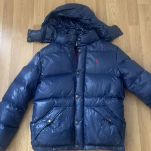 Blå dunjacka från Polo Ralph Lauren - Snygg mörkblå dunjacka från Polo Ralph Lauren med avtagbar huva och röd logga på bröstet. Jackan har stora fickor med tryckknappar, dragkedja och knappar framtill. Perfekt för kalla dagar när du vill hålla stilen.