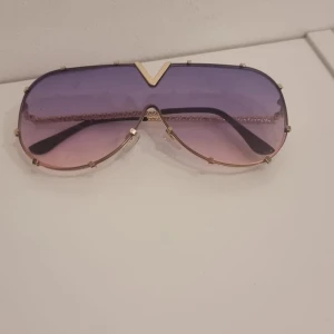 Lila-rosa solglasögon med gulddetaljer - Stora solglasögon med cool aviatorform och lila till rosa tonade glas. Guldfärgad metallram med dekorativa detaljer och en tydlig V-formad gulddetalj upptill. Perfekta för att sticka ut i mängden och ge en trendig vibe.