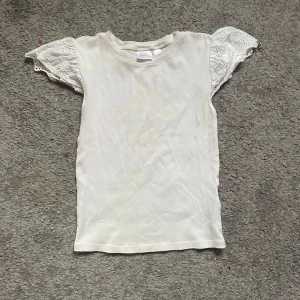 Vit blus med spetsärmar från Gina Tricot - Supersöt vit blus från Gina Tricot med ribbad kropp och korta, puffiga spetsärmar med broderade detaljer. Perfekt för dig som gillar en romantisk och trendig look. Rund halsringning och mjukt bomullsmaterial gör den extra bekväm.