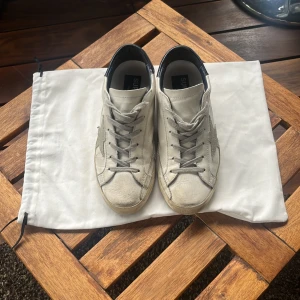 Golden Goose vita sneakers med stjärna - Golden Goose sneakers i vitt skinn med grå stjärndetalj på sidan och svarta hälpartier. Klassisk låg modell med snörning och platt sula. Snyggt slitna detaljer och ikonisk design som passar till allt. Kommer med originalpåse.