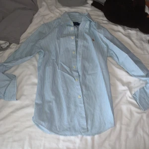 Randig skjorta från Ralph Lauren - Snygg blå- och vitrandig skjorta från Ralph Lauren med klassisk krage och broderad logga på bröstet. Skjortan har lång ärm, knappar framtill och är i bomull. Perfekt för dig som gillar stilrena och tidlösa plagg.