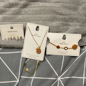 Smyckeset Smile Joyas halsband, armband, örhängen - Set med guldpläterat halsband med droppformad brun/orange sten, matchande armband med cirkulära stenar och flera par örhängen i pärl- och kristalldesign. Alla smycken är i rostfritt stål och har eleganta detaljer i guld och vitt.