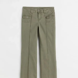 Svarta bootcut byxor med fickor - Snygga svarta bootcut byxor med markerade pressveck och två fickor framtill med lock och silverknappar. Byxorna har låg midja och är tillverkade i ett mjukt bomullsmaterial. Perfekta för en trendig och avslappnad look.