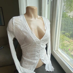 Vit blus i spets och knytband - Superfin vit blus i transparent mesh med broderade spetsdetaljer och v-ringning. Långa ärmar, knappar framtill och knytband i midjan för extra snygg passform. Perfekt för dig som gillar romantisk och trendig stil.
