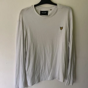 Vit långärmad tröja från Lyle & Scott - Vit långärmad tröja från Lyle & Scott med klassisk rund hals och den ikoniska gula fågel-loggan broderad på bröstet. Tröjan är i mjuk bomull och har en clean, stilren look som passar perfekt till jeans eller chinos.