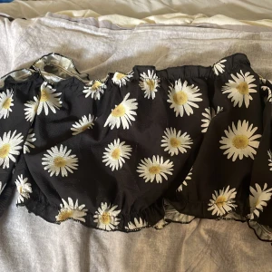 Svart offshoulder topp med prästkragar - Superfin svart offshoulder topp med stora vita prästkragar och gula detaljer. Toppen har resår vid axlar och midja för en snygg passform och är tillverkad i ett lätt, luftigt tyg. Perfekt för dig som vill sticka ut med ett blommigt mönster och somrig vibe.