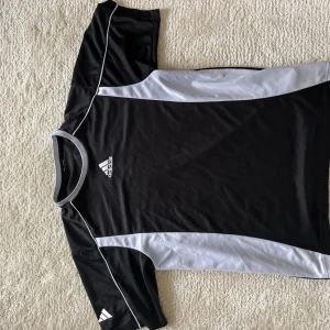 Svart och vit Adidas fotbollströja - Snygg svart fotbollströja från Adidas med vita detaljer längs sidorna och runt kragen. Klassisk Adidas-logga på bröstet och ärmen. Tröjan är kortärmad och tillverkad i ett lätt och ventilerande material, perfekt för träning eller match.