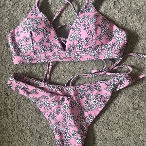 Rosa blommig bikini med snörning - Superfin bikini i rosa med vitt blommönster och svarta konturer. Toppen har trekantsform och snygg snörning i ryggen som ger en cool detalj. Underdelen är klassisk med samma blommiga print. Perfekt för stranden eller poolen! Oanvänd såklart 
