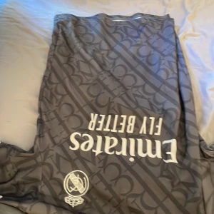 Real Madrid Vini Jr. 7 svart matchtröja - Säljer en svart Real Madrid fotbollströja med Vini Jr. och nummer 7 på ryggen. Tröjan har korta ärmar, mönstrad design med klubbens emblem och sponsortryck framtill. Tillverkad i lätt och ventilerande material, perfekt för dig som älskar fotboll och vill sticka ut.