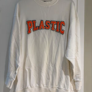 Vit sweatshirt från Madlady med tryck - Vit oversized sweatshirt från Madlady med stort broderat PLASTIC-tryck i orange och svart på bröstet. Tröjan har rund hals, långa ärmar och ribbade muddar. Perfekt för en chill och trendig look.