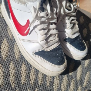 Nike Court Vision Low i vitt och rött - Nike Court Vision Low sneakers i vitt skinn med röda swoosh-detaljer och mörkblå partier vid tån. Klassisk retrostil med snörning och perforerad tåbox. Snygg kontrast mellan färgerna och en platt sula med rund tå. Perfekta för dig som gillar sportig streetstyle.