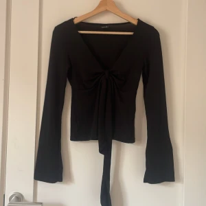 Svart långärmad topp med knyt - Svart långärmad topp med v-ringning och snygg knytning och öppet framtill. Ärmarna är lätt utsvängda och toppen har en figurnära passform. Perfekt att styla med jeans eller kjol för en trendig look.