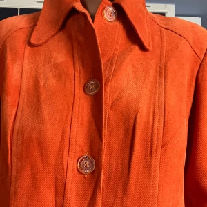 Orange manchesterkappa med stora knappar - Snygg vintage  orange kappa i manchester med  dekorativa knappar och två stora fickor framtill. Jackan har klassisk krage och raka ärmar, perfekt för hösten eller som en sensommar jacka. sticka u. Lång modell, räcker mig som är 163 över knäna precis. Fint skick förutom små kajal märken. 