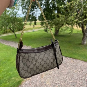 Gucci axelväska med GG-mönster - Snygg axelväska från Gucci i beige och brunt med klassiskt GG-mönster. Väskan har guldfärgad kedja, bruna skinn-detaljer och dragkedja upptill. Kommer med extra axelrem i brunt skinn. Perfekt storlek för det viktigaste och riktigt trendig look.