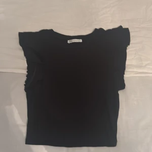 Svart volangärmad topp från Zara - Svart topp från Zara med snygga volangärmar och rund halsringning. Modellen är ärmlös med breda axelpartier och lätt rynkade sidor. Tunt, stretchigt tyg som känns skönt mot huden. Perfekt för dig som gillar stilrena och trendiga plagg.