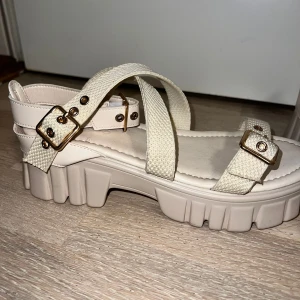 Chunky beige sandaler med spännen - Snygga chunky sandaler i beige med grov sula och breda textilband. Guldiga spännen och detaljer ger en cool touch. Remmarna korsas över foten och framtill, vilket ger en trendig look. Perfekta för dig som gillar statement-skor och vill sticka ut.