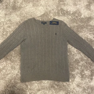Grå kabelstickad tröja Polo Ralph Lauren - Snygg grå kabelstickad tröja från Polo Ralph Lauren med klassisk rund halsringning och diskret broderad logga på bröstet. Tröjan har långa ärmar och ribbade muddar för en stilren look. Ny skick
