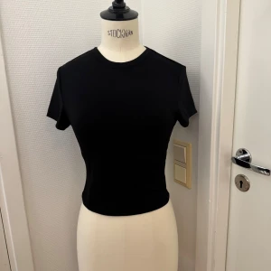 Svart topp med kort ärm XS - Svart stretch topp i XS med rund halsringning och korta ärmar. Jättemjuk. Nyskick! Sitter tight och har en enkel, clean look som passar perfekt till jeans eller kjol. Gjord i mjukt material som känns skönt mot huden.