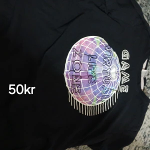 Svart t-shirt med färgglatt tryck - Cool svart t-shirt med ett stort färgglatt tryck framtill som föreställer en virtuell zon med texten 'GAME VIRTUAL LIFE ZONE'. T-shirten har rund hals och korta ärmar, perfekt för en avslappnad och trendig look.