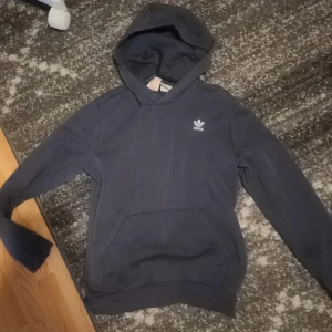 Adidas hoodie - Säljer en blå hoodie från Adidas med klassisk logga på bröstet. Tröjan har huva, magficka och är tillverkad i mjuk bomullsblandning. Perfekt för chill dagar eller när du vill ha en enkel och stilren look. Som Storlek XXS- liten s. Unisex. Rekommenderas bäst till xs