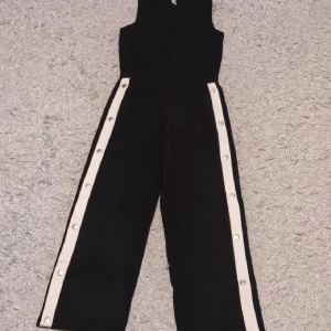 Svart byxdress med vita knappar - Snygg svart byxdress med breda ben och vita ränder längs sidorna, dekorerade med silvriga tryckknappar. Ärmlös modell med hög hals och relaxed fit. Perfekt för dig som vill sticka ut med en sportig och trendig look.