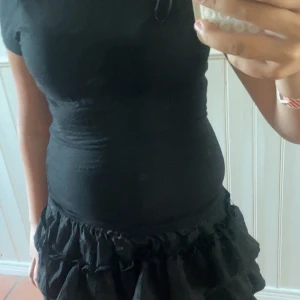 Svart kort klänning med rosett - Supersöt svart t-shirt och snygg öppen rygg med knytband i satin. T-shortsen har korta ärmar och figurnära passform, perfekt för dig som gillar en stilren look med en twist. Materialet känns mjukt och följsamt. Använd ca 2-3 gånger