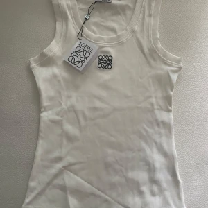 Vit ribbad linnetopp från Loewe - Supersnygg vit ribbad linnetopp från Loewe med broderad svart logga på bröstet. Tidlös och clean design med rund halsringning och ärmlös passform. Perfekt att styla med jeans eller kjol för en fräsch look.