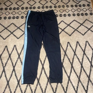 Svarta träningsbyxor från Lacoste - Snygga svarta träningsbyxor från Lacoste med ljusblå sidopanel och broderad logga på låret. Byxorna har resår i midjan och vid bensluten för en skön passform. Perfekta för sport eller chill, med klassisk streetkänsla och sportig look.