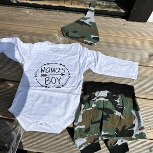 Vit långärmad body med tryck - Vit långärmad body med svart text 'Mama's Boy' och pilmotiv framtill. Bodyn har rund hals och tryckknappar i grenen för enkel påklädning. Perfekt för en cool och bekväm stil till de minsta. Cirka för 1 år +