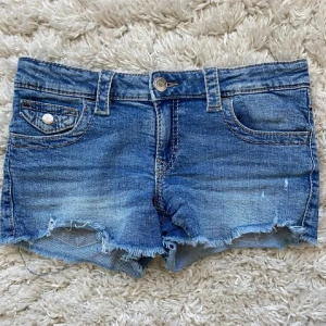 Blå jeansshorts med rå kant - Snygga blå jeansshorts med slitna detaljer och råa kanter. Klassisk femficksmodell med knapp och dragkedja framtill samt fickor med lock och knapp bak. Perfekta för varma dagar och en avslappnad look.