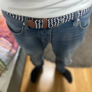 Randigt flätat bälte i blått och vitt - Snyggt flätat bälte med randigt mönster i marinblått och vitt. Bältet har detaljer i brunt skinn vid spännet och är perfekt för att ge jeansen en cool touch. Passar dig som gillar stilrena accessoarer med lite edge. Helt nytt!