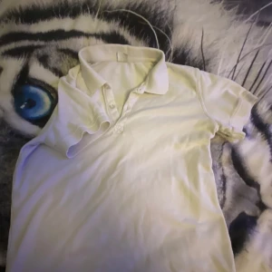 Vit pikétröja från Lindex - Klassisk vit pikétröja från Lindex med krage och knappar framtill. Tröjan har korta ärmar och är tillverkad i mjuk bomull, perfekt för en clean och enkel stil. Passar dig som gillar basic och tidlösa plagg.