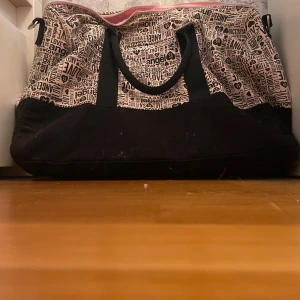 Victoria's Secret weekendbag  - Stor och rymlig weekendbag från Victoria's Secret med svart och ljusrosa färg. Väskan har coolt textmönster med logga och hjärtan, svarta handtag och rosa insida. Perfekt för resor eller gymmet, med dragkedja upptill och mjukt material. Drag kedjan är lite trasig tyvärr men man kan fortfarande använda väskan❤️