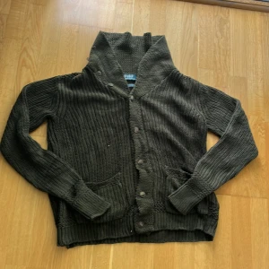 Mörkgrå stickad kofta med krage - Säljer en mörkgrå stickad kofta från Polo Ralph Lauren med hög krage och knäppning framtill. Koftan har två fickor och ribbade muddar vid ärmslut och nederkant. Perfekt för lager-på-lager och chill höst- och vinterstil.