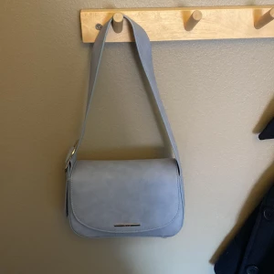 Ljusblå axelväska i mocka-look - Säljer en stilren ljusblå axelväska från Bershka mocka-liknande material. Väskan har ett enkelt, clean design med silverfärgad detalj framtill och magnetlås. Invändigt finns ett fack med dragkedja för småsaker. Perfekt för dig som gillar minimalistisk stil. Finns tyvärr två små fläckar en på insidan av väska och en lite på ”locket”.❤️🥰 pris kan diskuteras💵