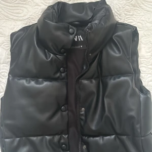 Svart väst Zara - Säljer en svart, quiltad dunväst från Zara i skinnimitation. Västen har hög krage, tryckknappar och dragkedja framtill. Skriv för mer info. Den är i perfekt skick