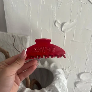 Röd hårklämma från Skims - Cool röd hårklämma från Skims med glittriga stenar som bildar loggan på ena sidan. Klämman är i plast och har en klassisk form med breda taggar som håller håret på plats. Perfekt accessoar för att lyfta din look med en pop av färg.