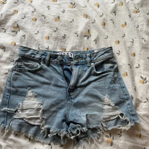 Blåa slitna jeansshorts från Stay - Snygga ljusblå jeansshorts från Stay med hög midja, slitna detaljer och fransig kant. Klassisk femficksmodell med dragkedja och knapp framtill. Perfekta för sommaren och ger en cool, avslappnad vibe.