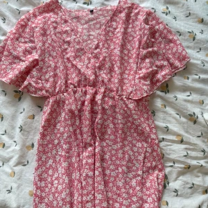 Rosa blommig kortklänning med v-ringning - Supersöt rosa kortklänning med vitt blommönster och v-ringning. Klänningen har korta, vida ärmar och resår i midjan för en skön passform. Perfekt för varma dagar och ger en fräsch, somrig vibe!