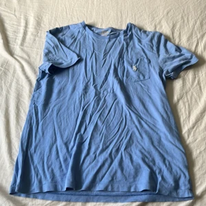 Blå t-shirt från Polo Ralph Lauren - Snygg ljusblå t-shirt från Polo Ralph Lauren i bomull. Modellen är slim fit och har en liten bröstficka med den klassiska broderade loggan. Perfekt basic-plagg med skön passform och stilren look.