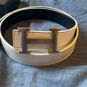 Vitt skärp med stor H-spänne - Snyggt vitt bälte med en stor, silverfärgad H-formad metallspänne. Bältet är gjort i präglat skinn och har svarta kanter på insidan. Perfekt accessoar för att lyfta din outfit med en clean och modern look.