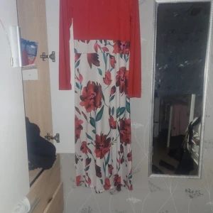 Långklänning med röd topp och blommig kjol - Snygg långklänning med enfärgad röd överdel och långa ärmar. Kjolen är vit med stora röda och gröna blommor som ger en färgstark och fräsch look. Perfekt för dig som vill sticka ut med en romantisk och trendig stil.