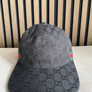 Svart keps från Gucci med GG-mönster - Snygg svart keps från Gucci med klassiskt GG-monogram över hela kepsen. Kepsen har gröna och röda ränder på sidorna och justerbar rem baktill. Tillverkad i bomull och polyester, perfekt för dig som vill ha en exklusiv streetstyle-look.