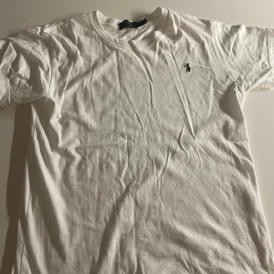 Vit t-shirt från Polo Ralph Lauren - Klassisk vit t-shirt från Polo Ralph Lauren med broderad logga på bröstet. T-shirten är i mjuk bomull, har rund hals och korta ärmar. Perfekt basic-plagg med stilren design och skön passform.