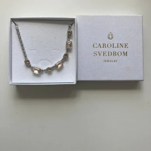 Caroline Svedbom halsband med kristaller - Supersnyggt halsband från Caroline Svedbom med stora ljusrosa och champagnefärgade kristaller på en silverfärgad kedja. Stenarna är både runda och rektangulära och ger ett riktigt lyxigt intryck. Perfekt för dig som vill sticka ut med lite extra bling.